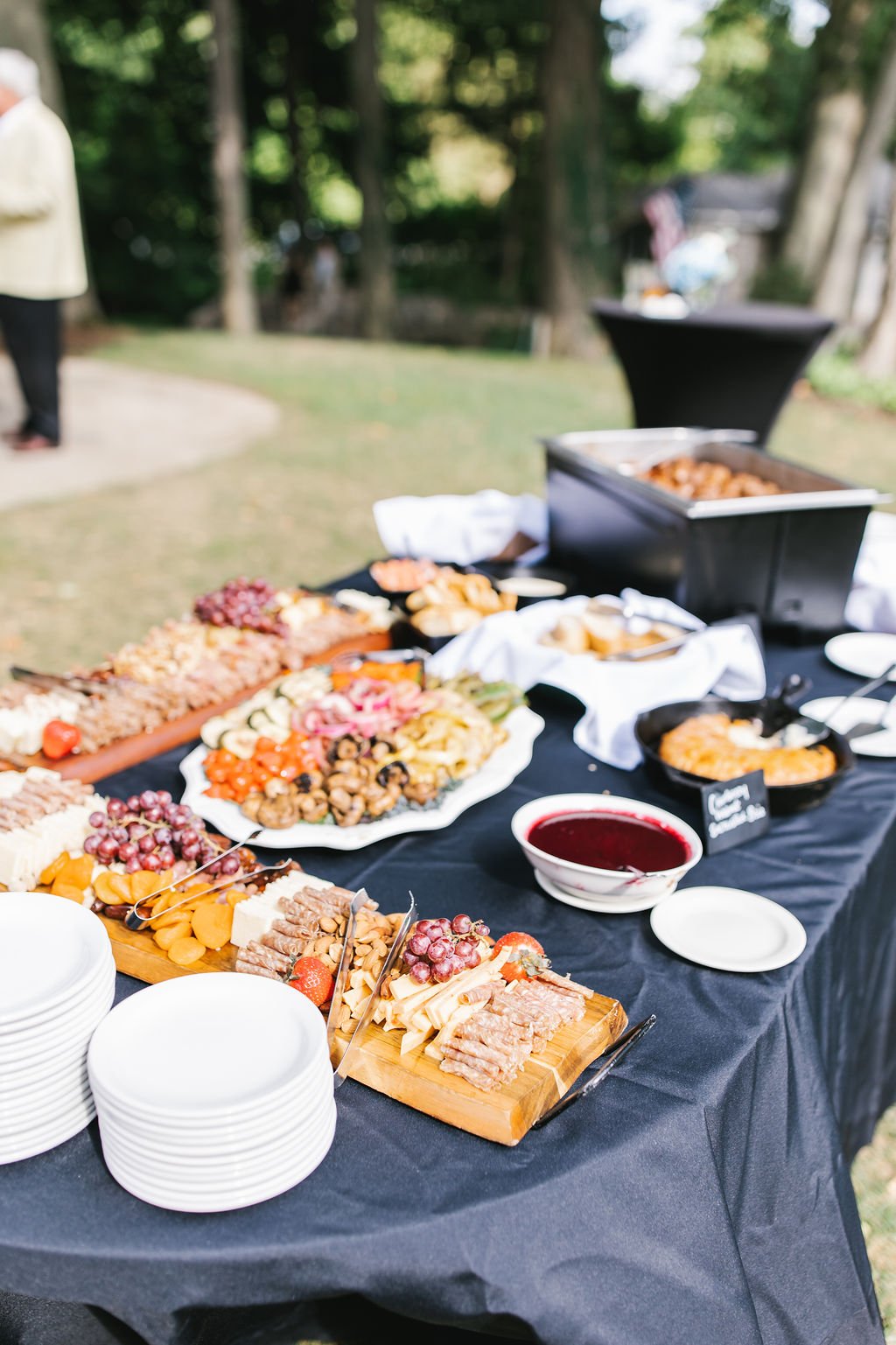 Outdoor buffet table with cheese, grapes, prosciutto, bread, and condiments on a black tablecloth, set in a green park with trees in the background.
