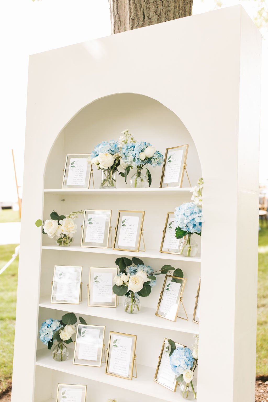 White wedding seating chart display with framed table assignments and floral decorations, featuring white and blue hydrangeas and white roses.