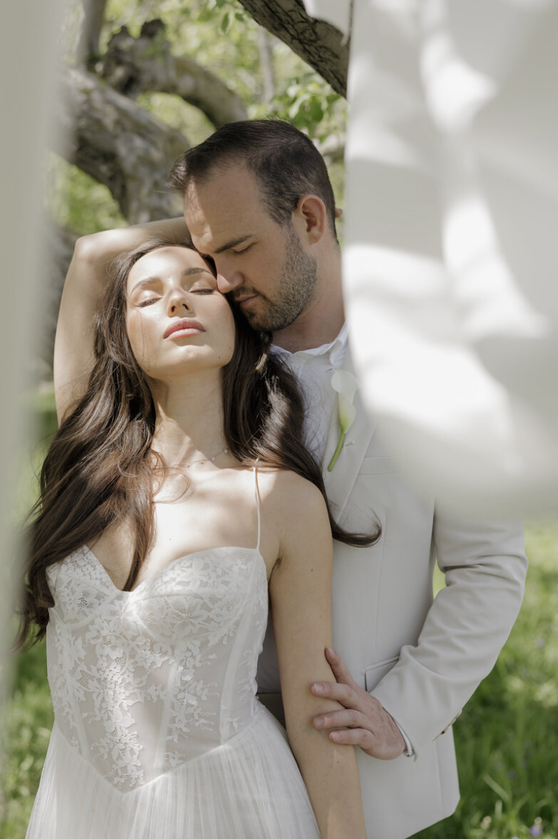 A bride and groom embrace outdoors under a tree, with sunlight filtering through leaves, sharing an intimate moment.