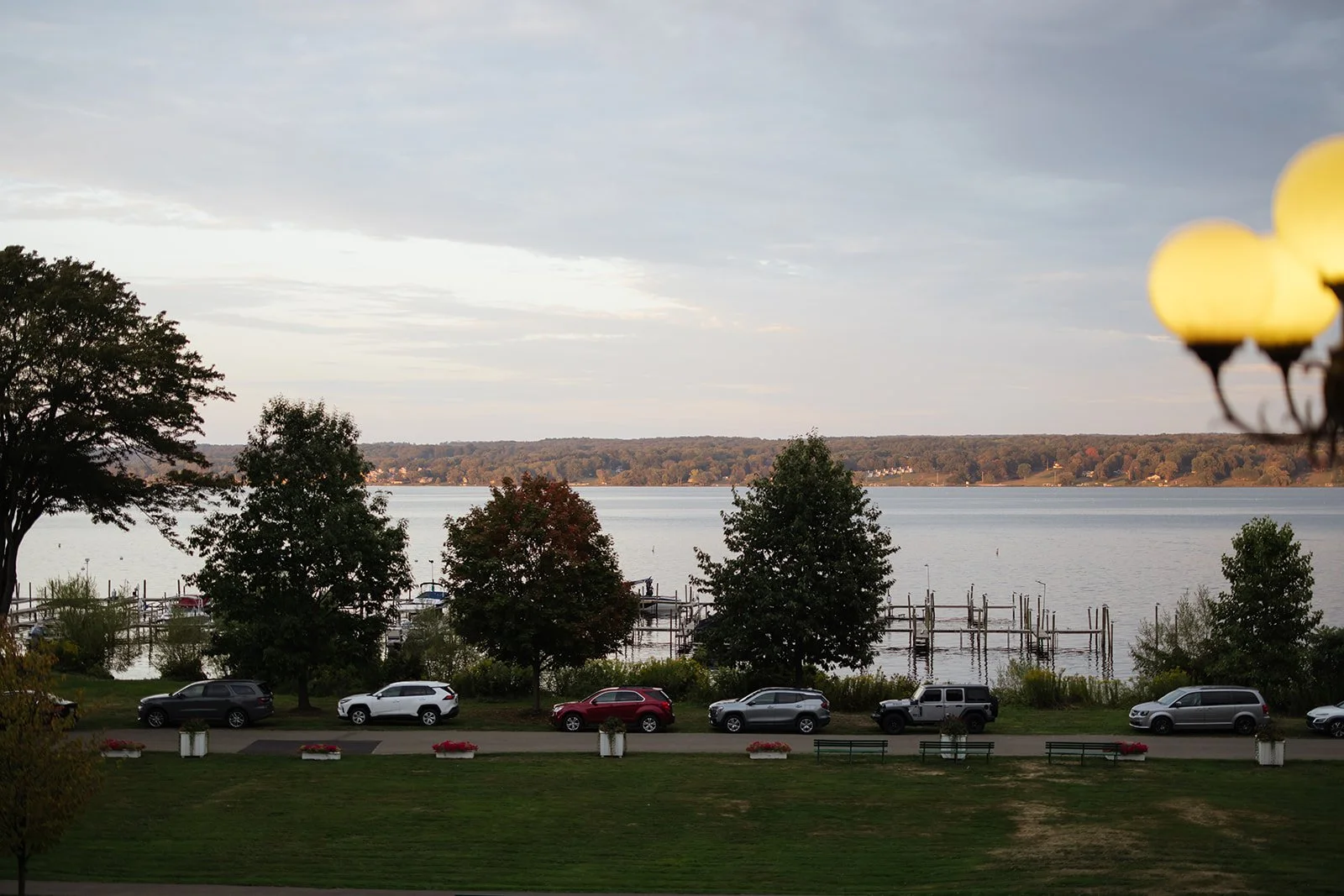 A lakeside scene with parked cars along a walkway, trees, a dock with boats, and a distant shoreline under a cloudy sky during late afternoon or early evening.