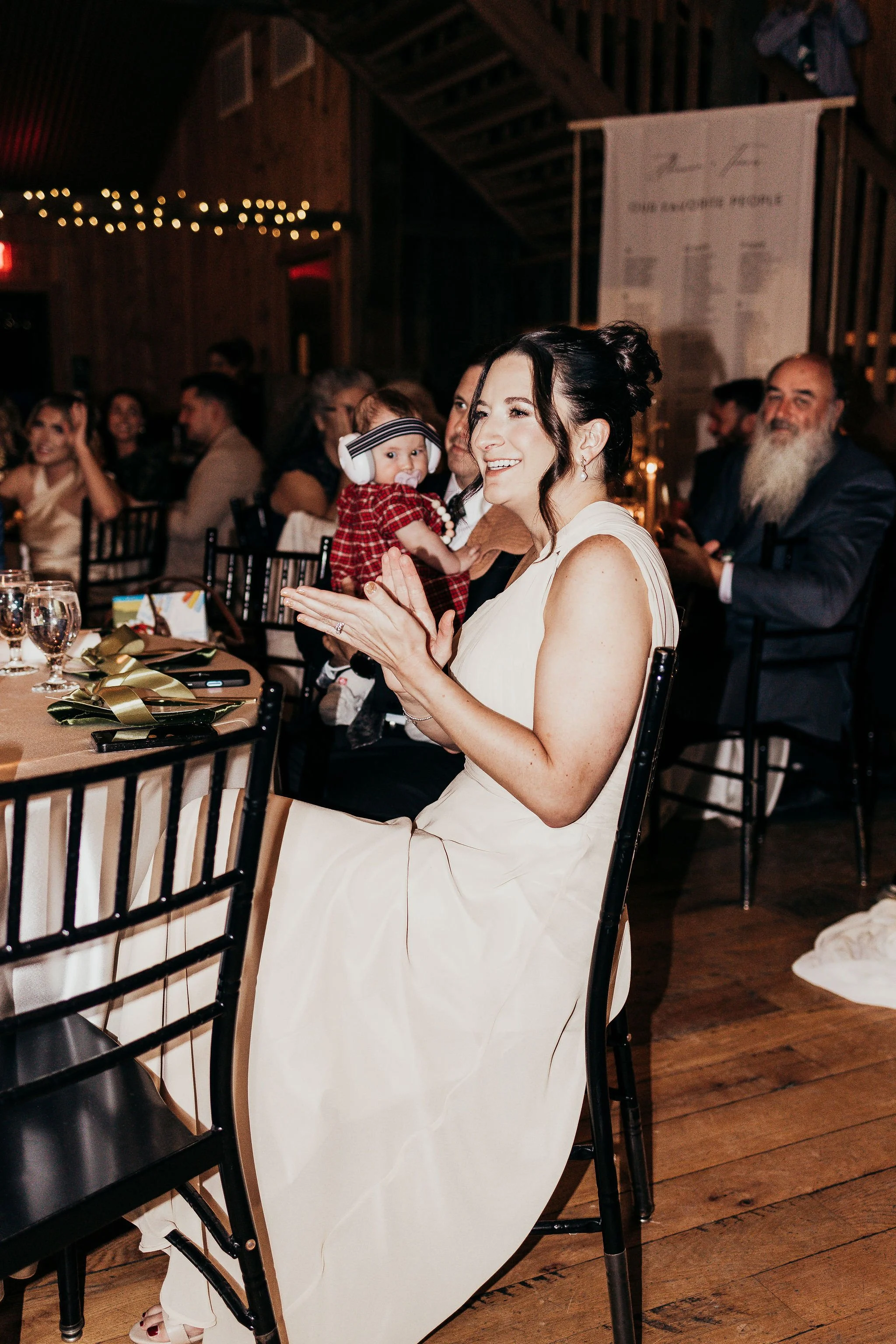 A woman in a white dress holding a baby with headphones at a dinner event, with other guests seated at tables in a wooden venue decorated with string lights.