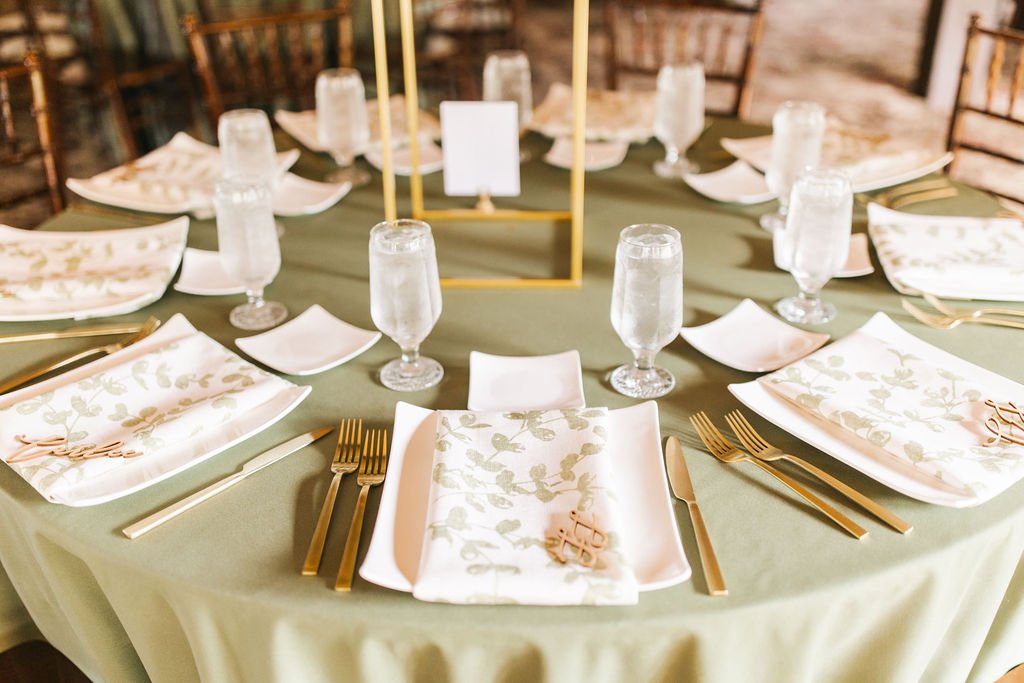 Elegant round table set with white and gold flatware, floral-patterned napkins, glasses of water, and a gold geometric centerpiece in a rustic setting.
