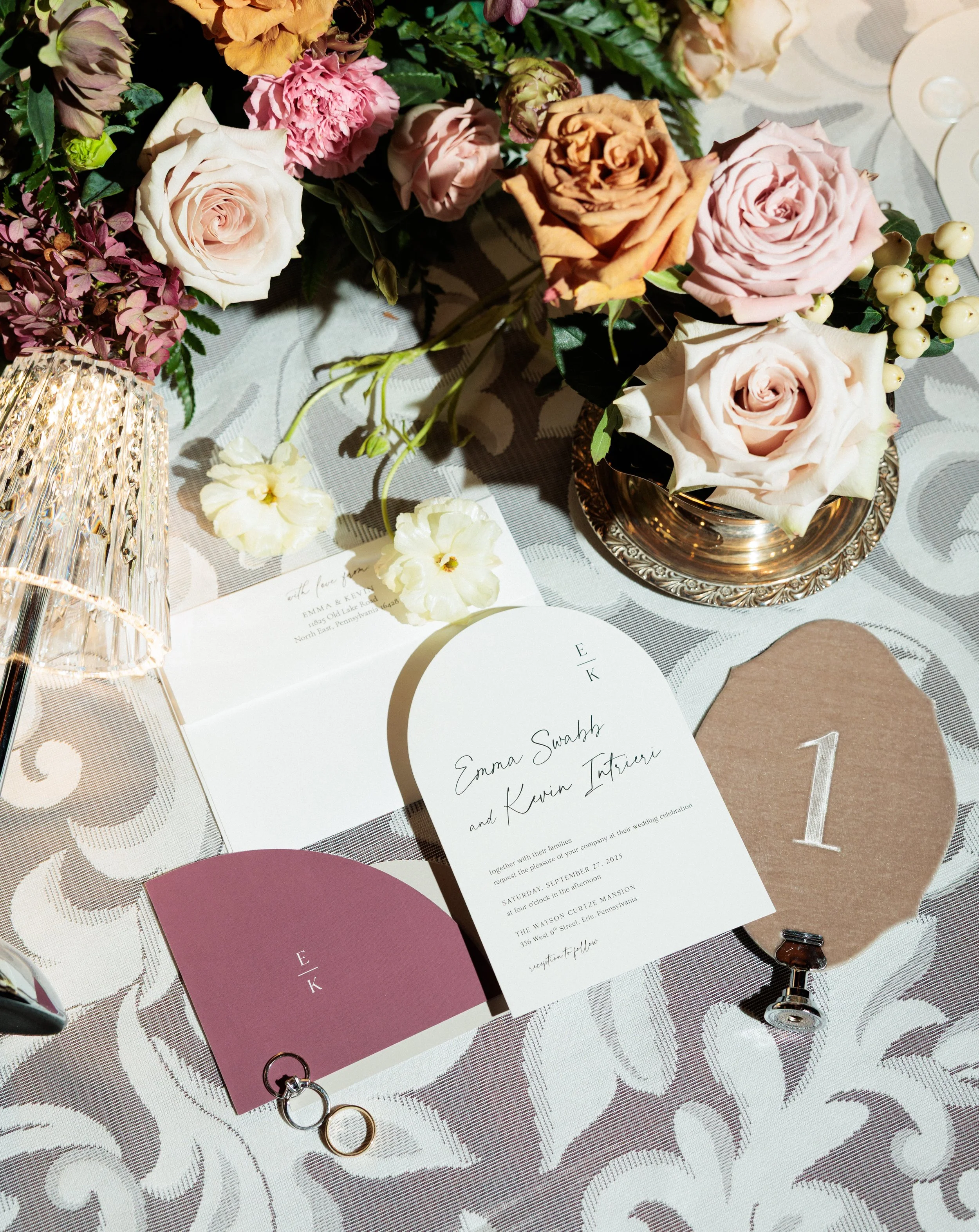 A wedding invitation card, wedding rings, and a decorative flower bouquet on a patterned tablecloth.