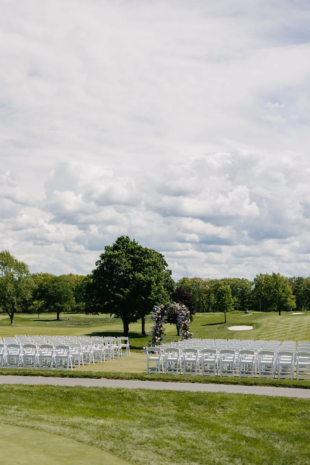 Outdoor wedding setup on a golf course with white chairs arranged in rows, an arch decorated with flowers, and trees under a partly cloudy sky.