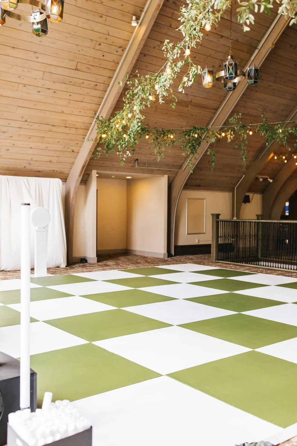 Event venue with a wooden arched ceiling, decorated with hanging greenery and string lights, with a checkered green and white dance floor.