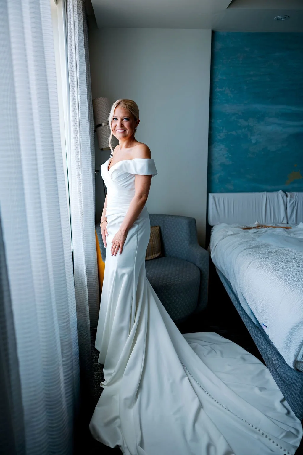 A bride in a white off-the-shoulder wedding gown standing near a window in a hotel room, smiling. The room has a bed and a blue accent wall.