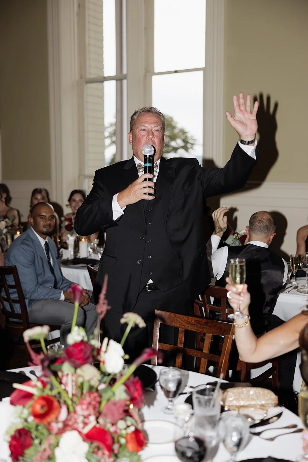 A man in a tuxedo speaking into a microphone at a formal event, with guests seated at tables decorated with flowers and candles, in a well-lit room with large windows.