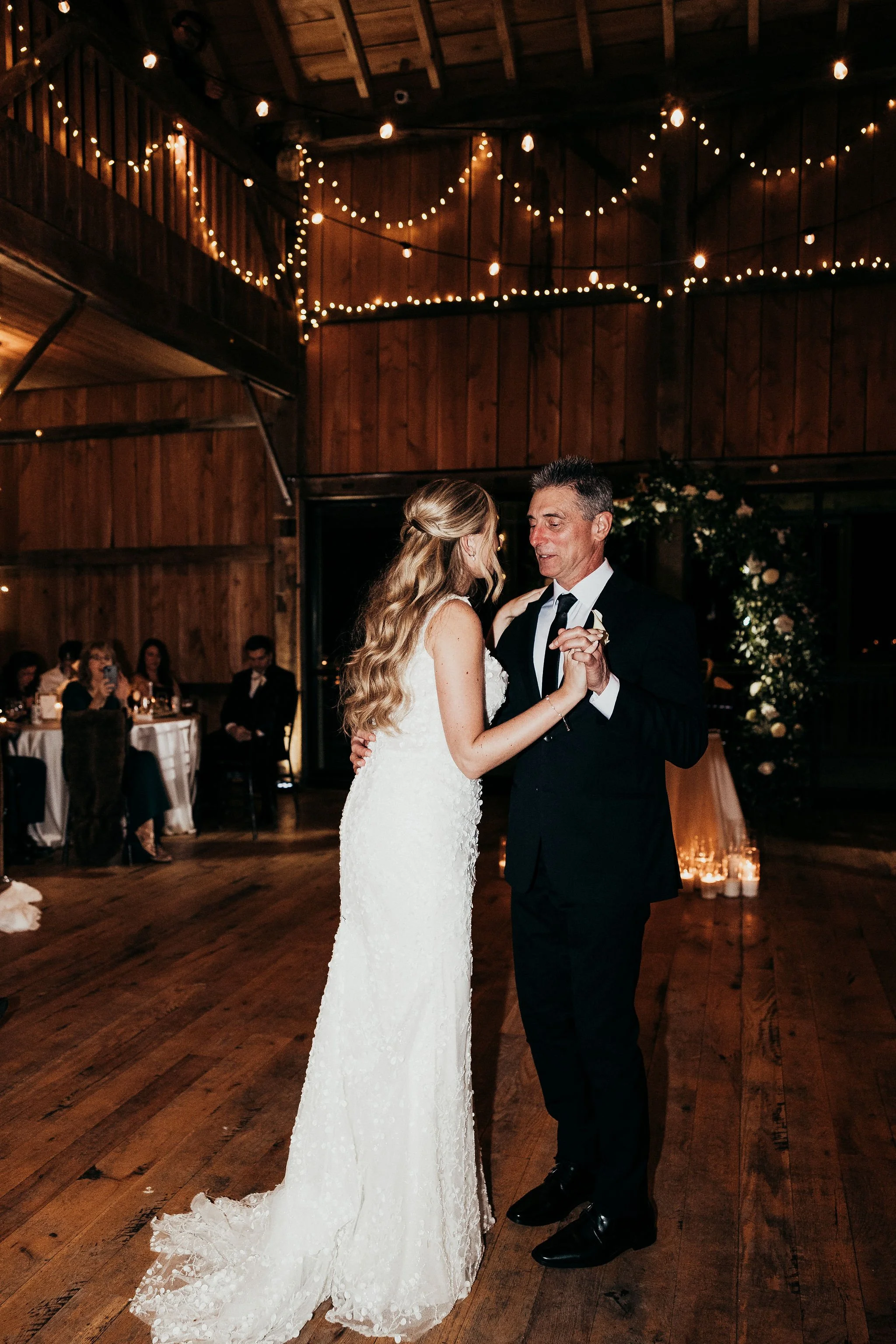A bride and groom are dancing at their wedding reception inside a wooden venue. The bride is wearing a white lace wedding gown, and the groom is wearing a black tuxedo with a white shirt and black tie. String lights and candles decorate the backgroun