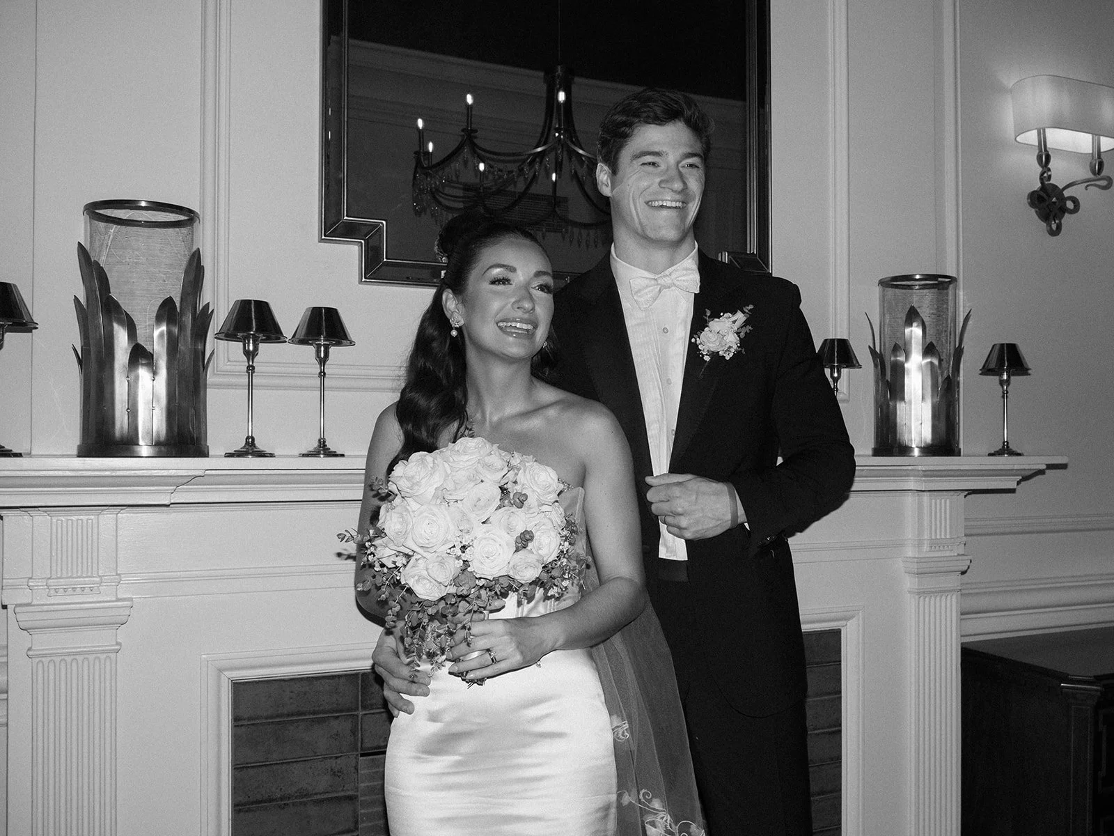Black and white photo of a newlywed couple smiling, standing in front of a fireplace. The bride holds a bouquet of roses, and the groom wears a tuxedo with a bow tie. A chandelier and decorative candles are reflected in a mirror above the fireplace.