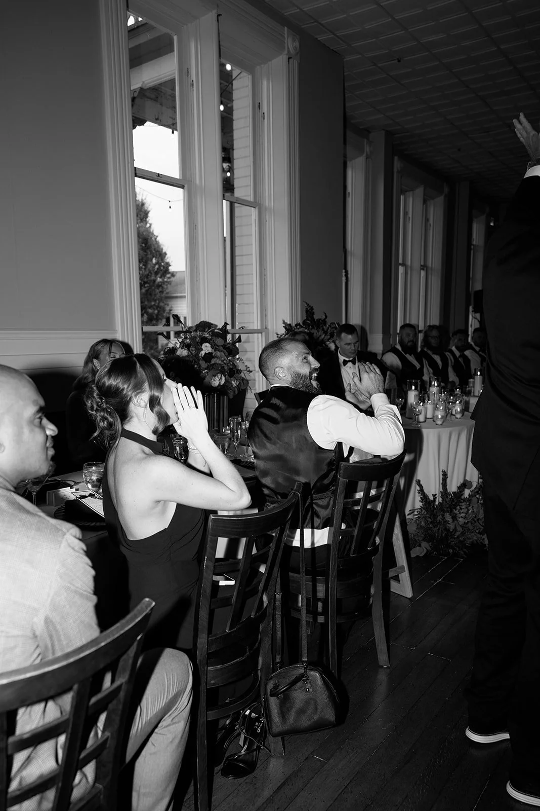 Guests at a formal event, seated at tables, some with hands over their faces, watching a person standing and speaking. The venue has large windows and floral arrangements on tables.