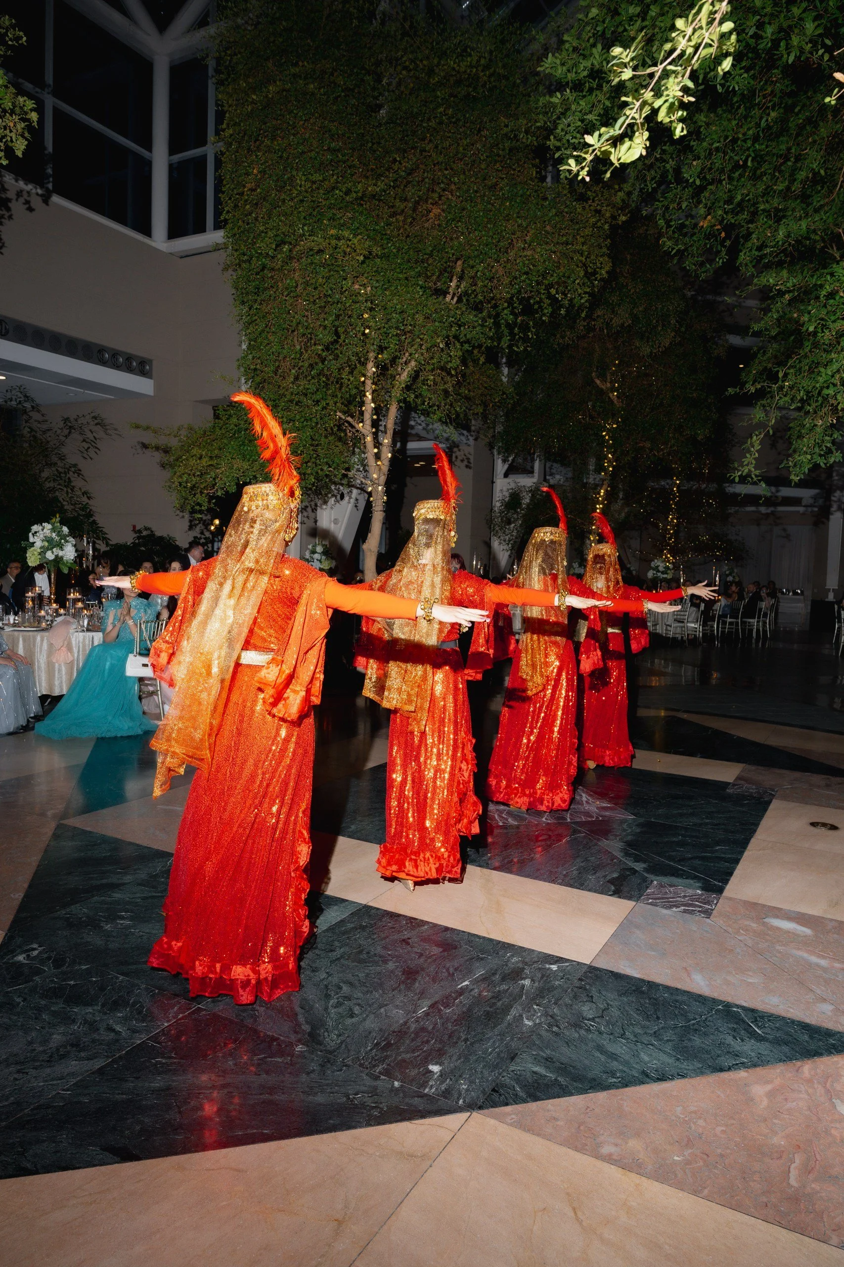 Women dressed in red and gold costumes with feathered headdresses are performing a traditional dance at a formal event or celebration in an indoor space with tables and guests seated around.