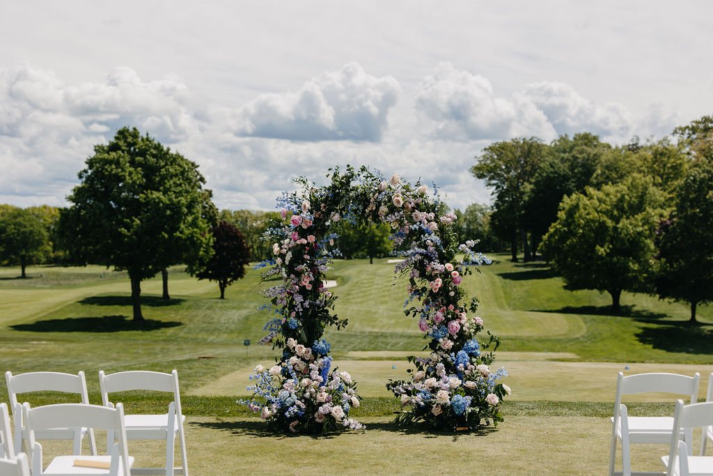 Wedding ceremony setup outdoors with a floral arch and white chairs on a grassy field, trees in the background under a partly cloudy sky.