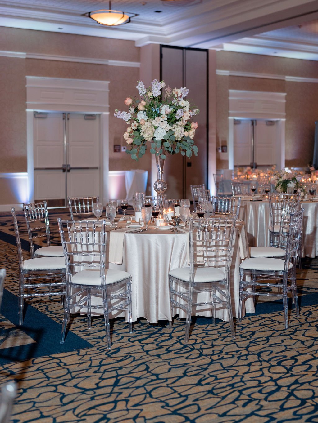 Elegant round table set for a wedding reception with a large floral centerpiece, white tablecloth, glassware, and clear chairs in a decorated banquet hall.