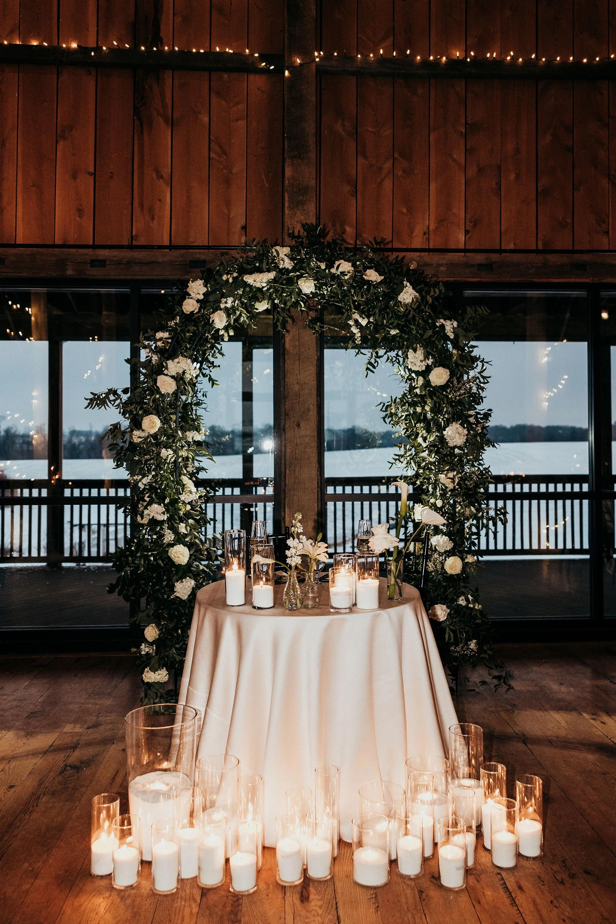 An indoor wedding or event setup with a round table covered with a beige tablecloth, decorated with numerous white candles in glass holders, and surrounded by a green floral arch with white flowers and leaves. Behind is a large window overlooking a b