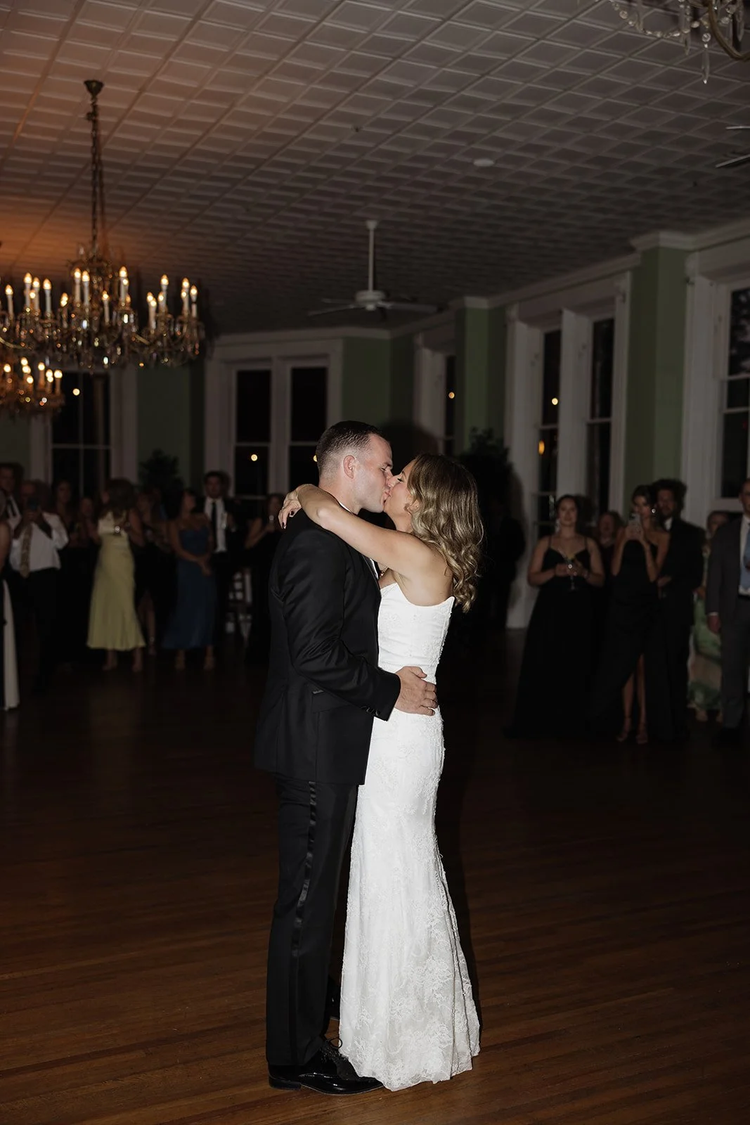 A bride and groom are sharing their first dance at their wedding reception, surrounded by guests in a large, elegantly decorated room.