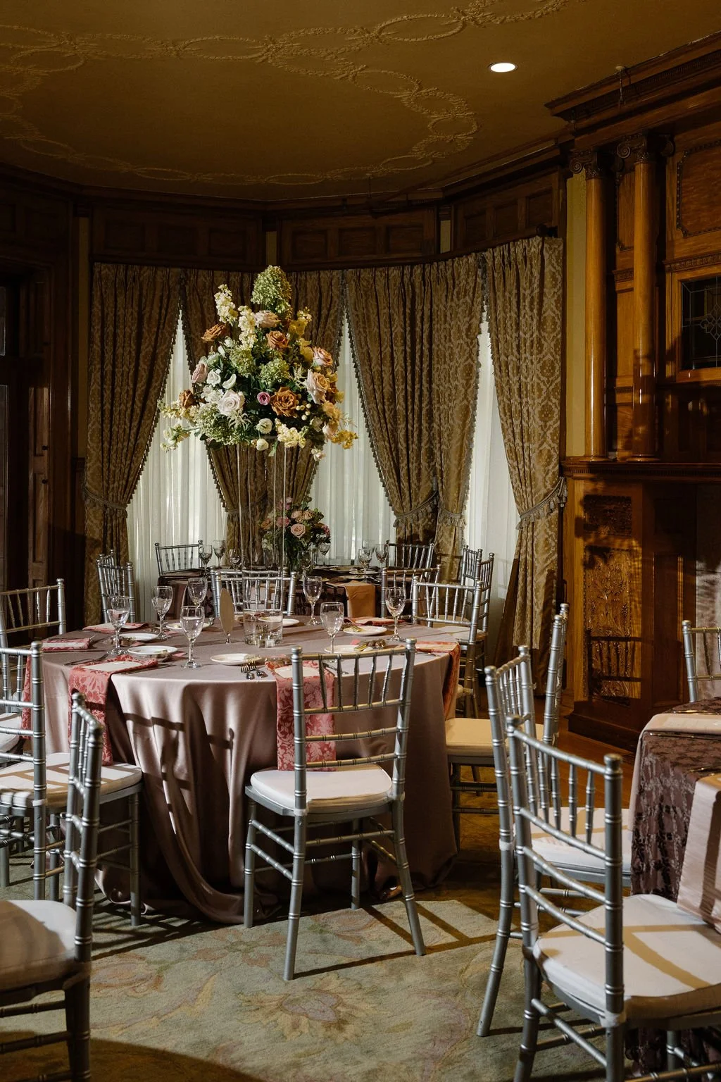 Elegant dining room with round table set with glasses and dishes, surrounded by silver chairs, large floral centerpiece, and gold patterned curtains with wood-paneled walls.