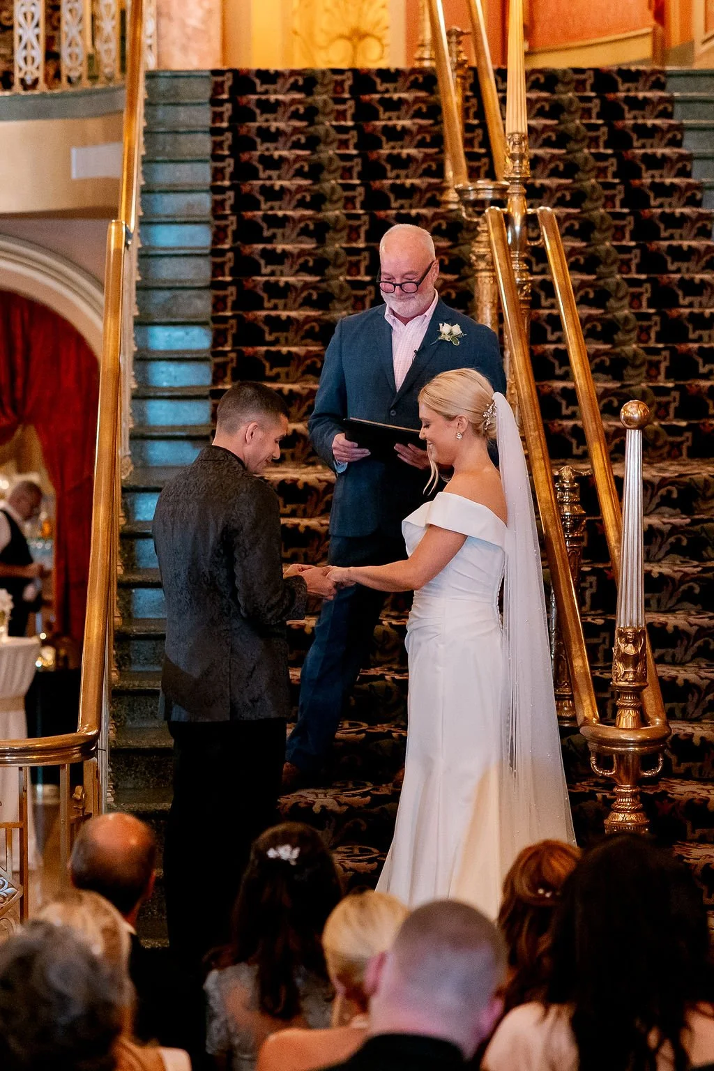 A wedding ceremony with a bride and groom exchanging vows on a staircase, with an officiant standing behind them, in a decorated venue.