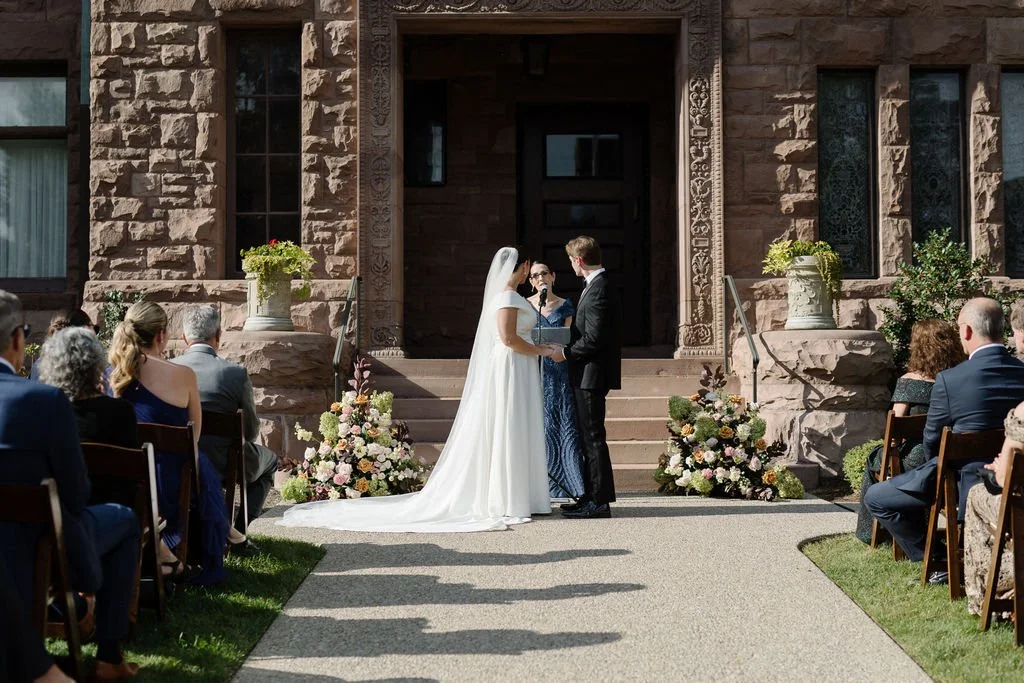 Bride and groom exchanging vows in a wedding ceremony outside a stone building, with guests seated on either side and floral arrangements at the steps.