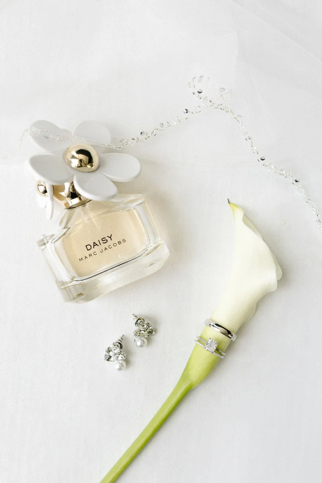 A bottle of Marc Jacobs Daisy perfume with white floral cap, a white calla lily flower with wedding rings on its stem, and a pair of pearl and diamond earrings on a white background.