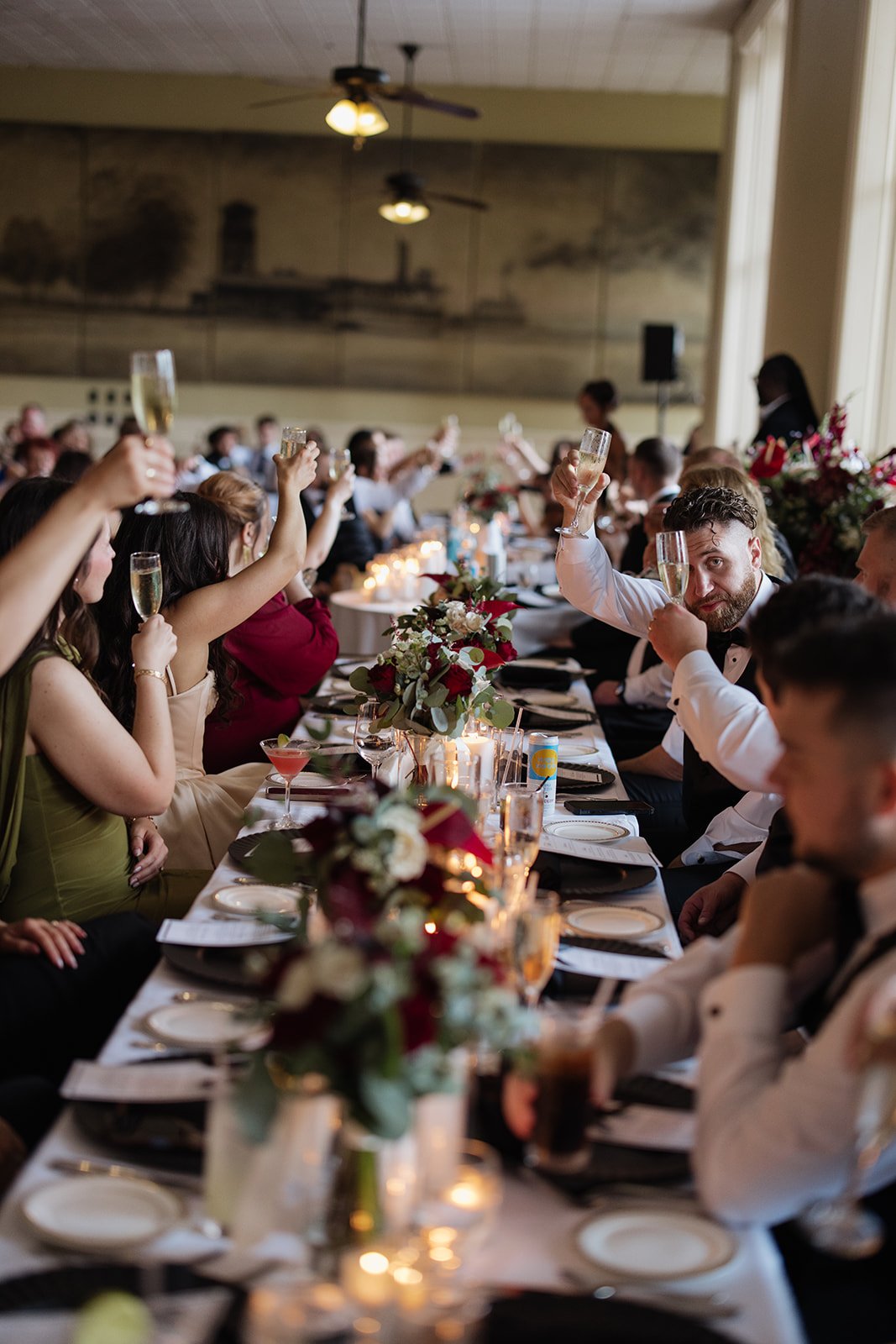 People sit at a long banquet table, raising champagne glasses in a toast, decorated with flowers and candles, at a formal celebration or wedding reception.