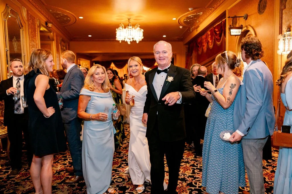 Guests at a formal wedding reception, including a bride in a white gown, a groom in a black tuxedo, and other attendees in elegant attire, inside a decorated banquet hall with chandelier lighting.