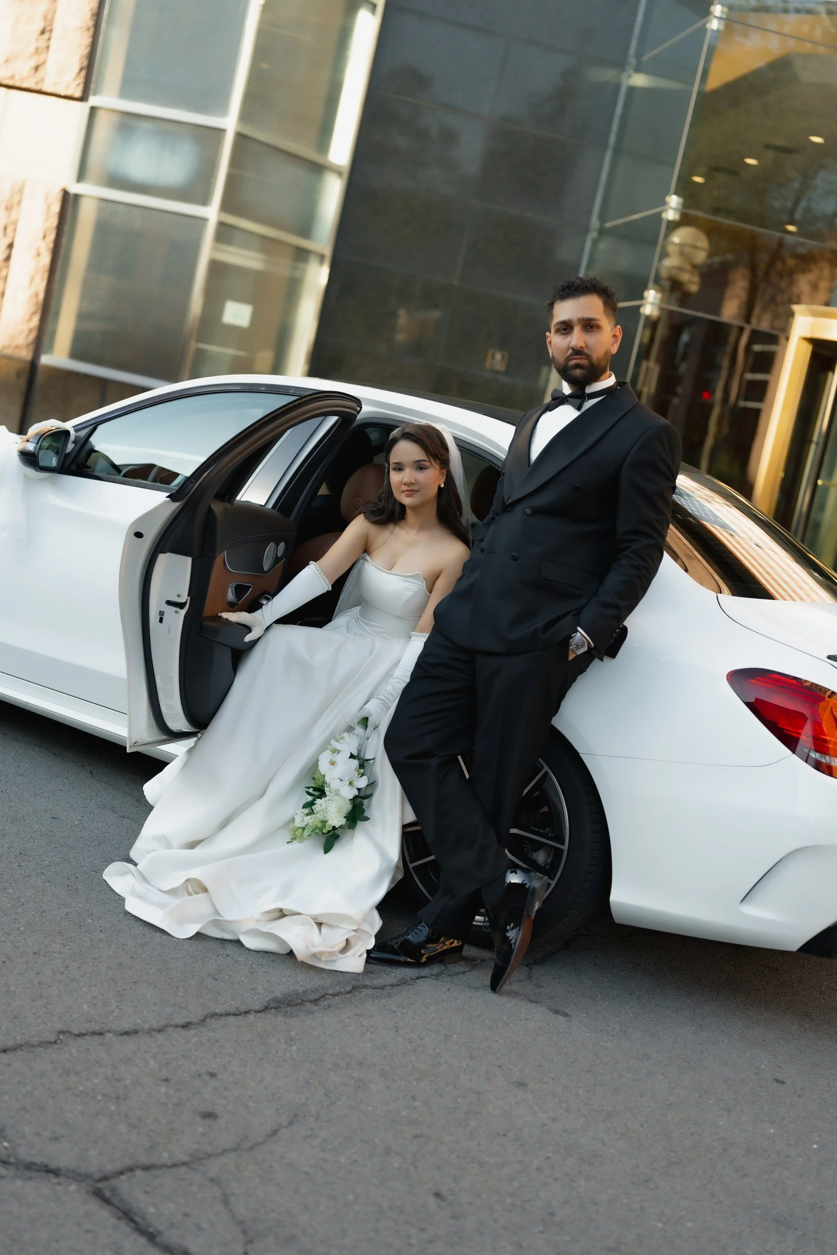 A newlywed couple with light skin posing by a white luxury car in front of a modern building. The bride, sitting in the car, is wearing a white wedding gown with long gloves and holding a bouquet of white flowers. The groom, standing beside her, is d