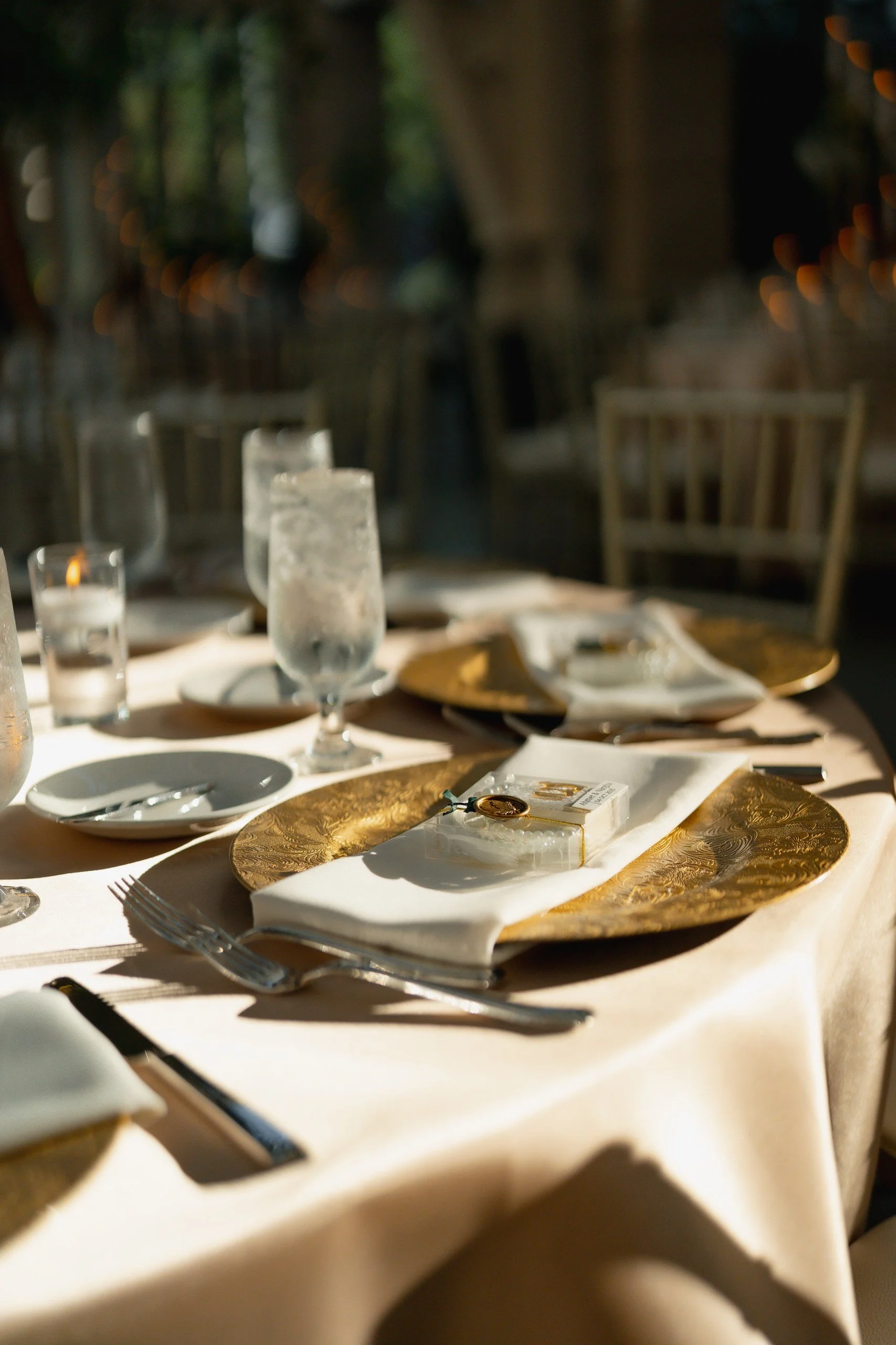 A tablescape set for an event, featuring gold chargers, white plates, napkins, silverware, glassware including a water pitcher and glasses, candles, and a small decorative item, with blurred background of chairs and trees.