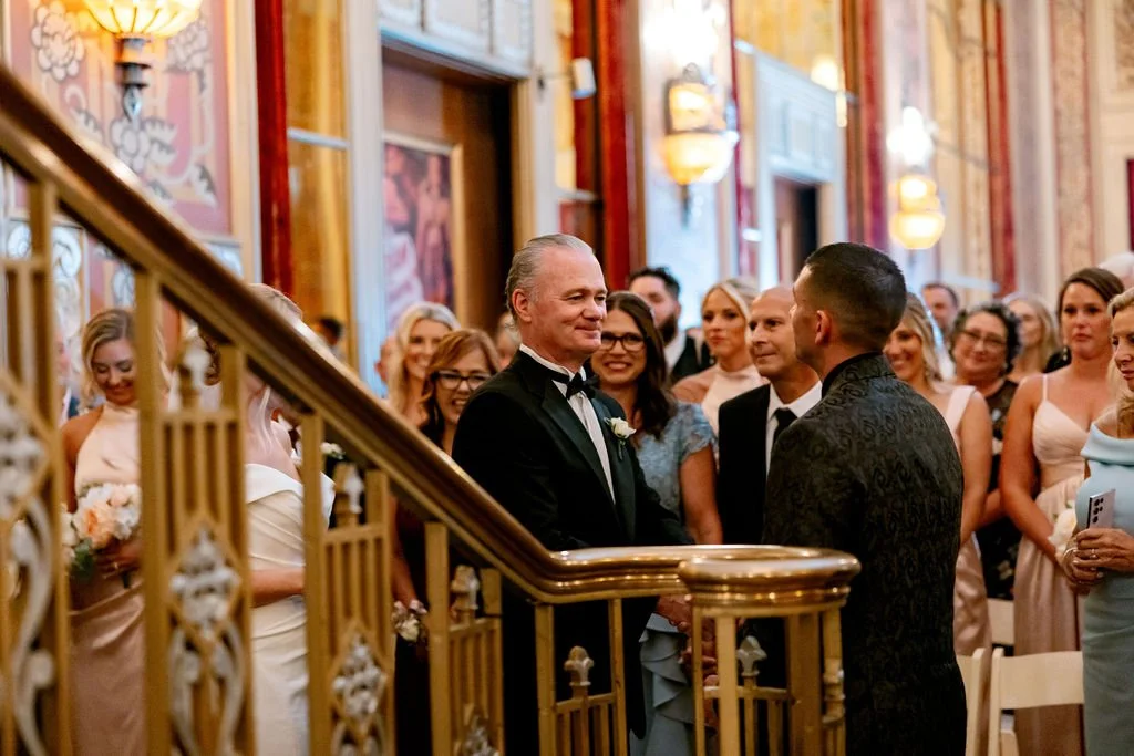 A wedding ceremony with a groom and officiant exchanging vows in front of an audience in an ornate indoor setting