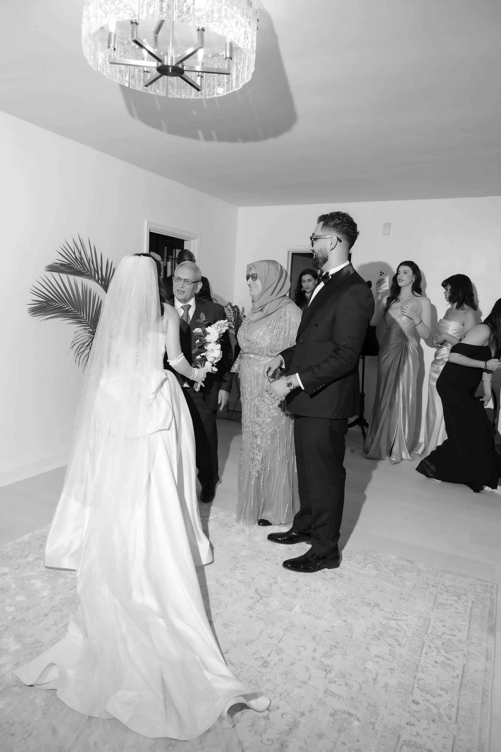 A group of people, including a bride in a wedding gown and veil, standing with an older man, a woman, and a groom in a tuxedo, at a wedding ceremony indoor with guests in elegant dresses watching.