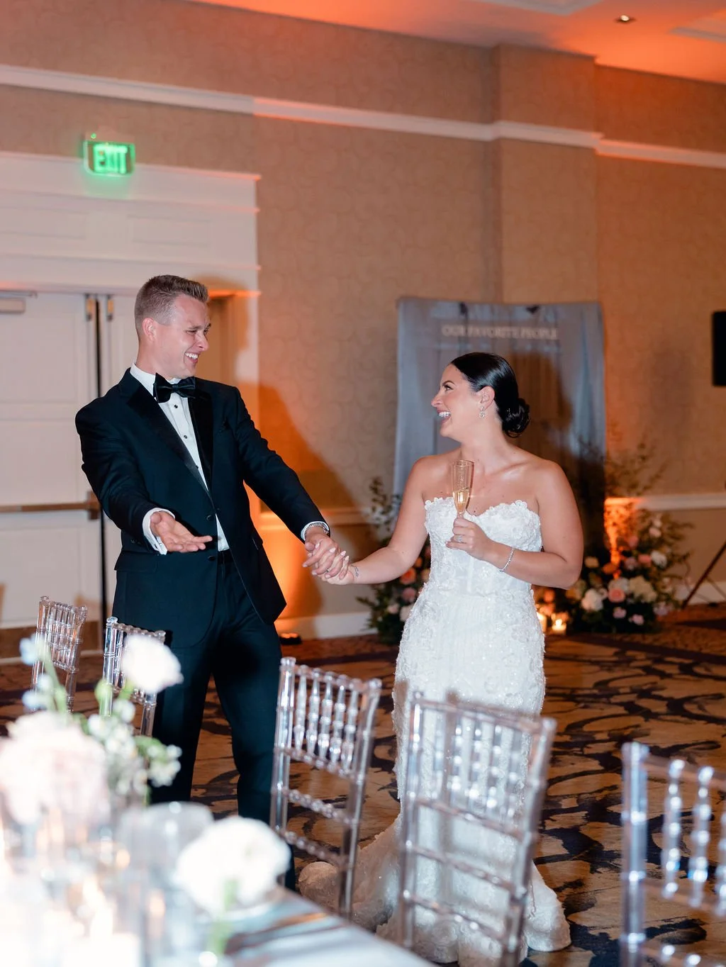 Bride and groom dancing and smiling at wedding reception, woman holding champagne glass, man in tuxedo.