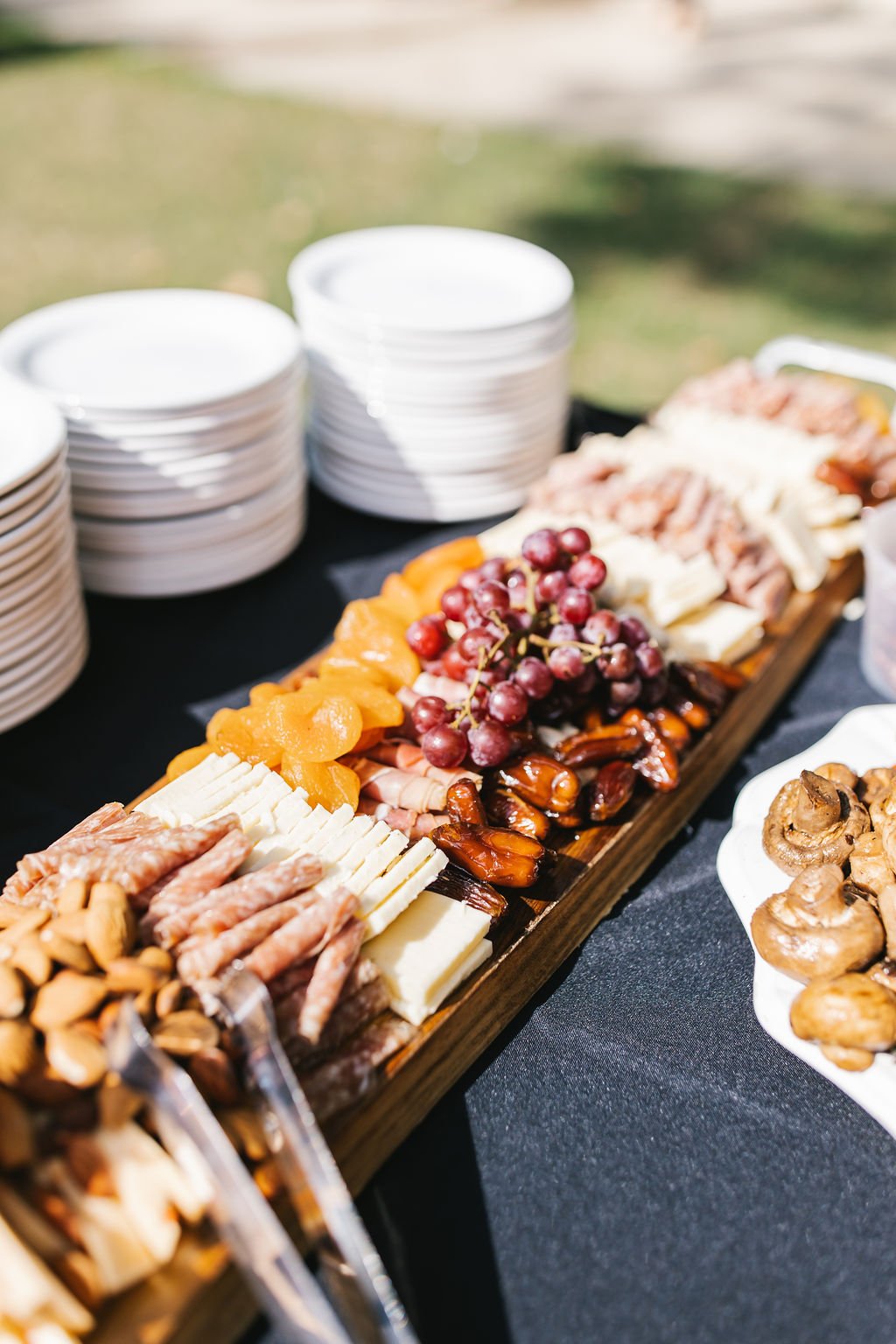 Assorted cheese, grapes, and charcuterie on a wooden board at an outdoor gathering.