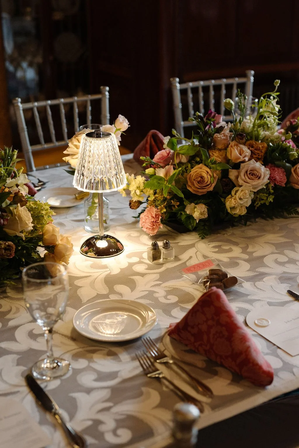 A decorated dining table with floral arrangements, a small table lamp, plates, silverware, a glass of water, and a napkin.