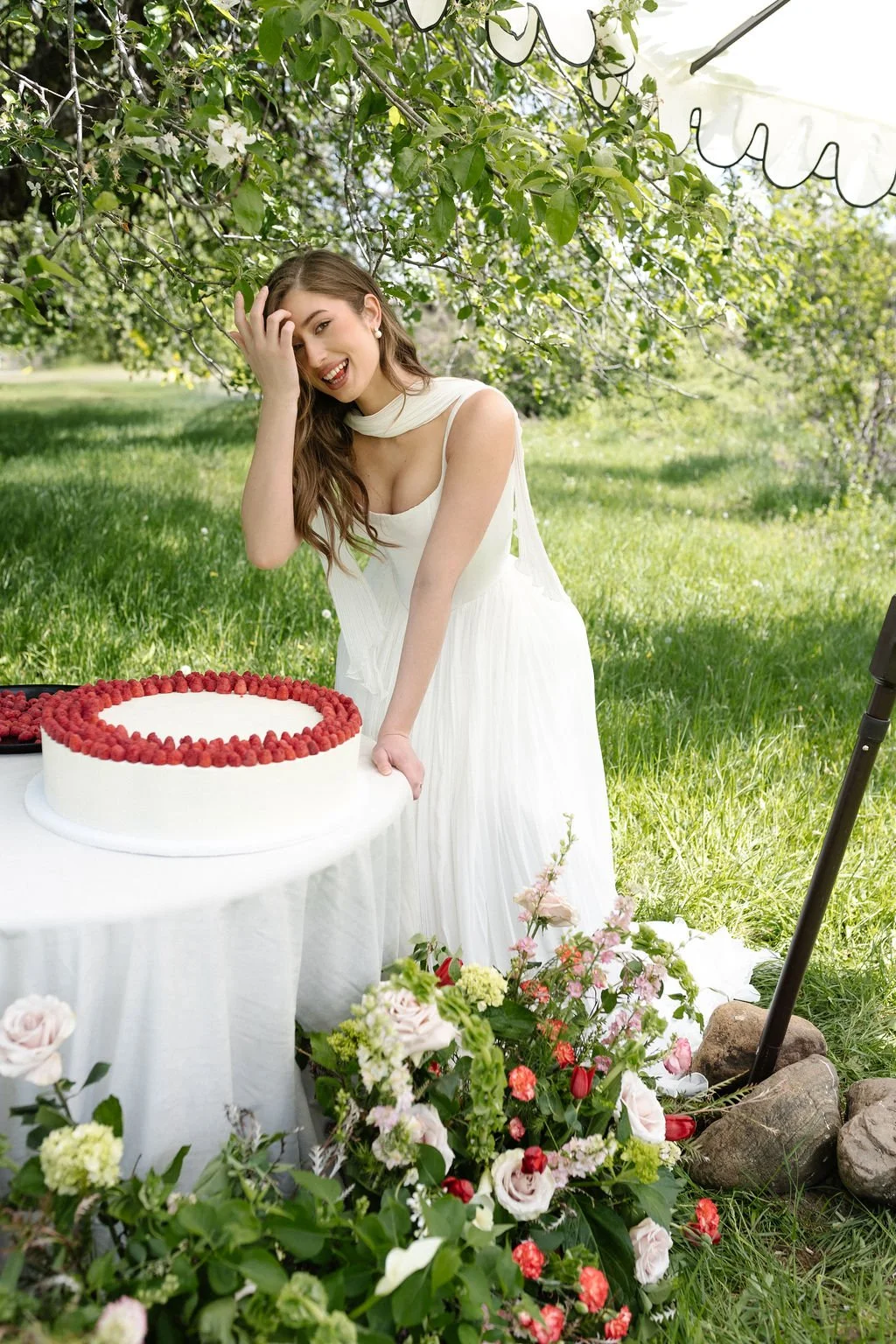 A young woman in a white dress happily poses with a large cake decorated with raspberries on a table outdoors under a tree during daytime. There are colorful flowers in front of the table and rocks on the ground nearby.