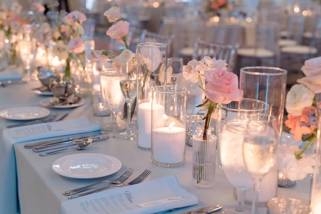 A beautifully set banquet table with white tablecloths, floral arrangements, and lit candles, prepared for a formal event or wedding reception.