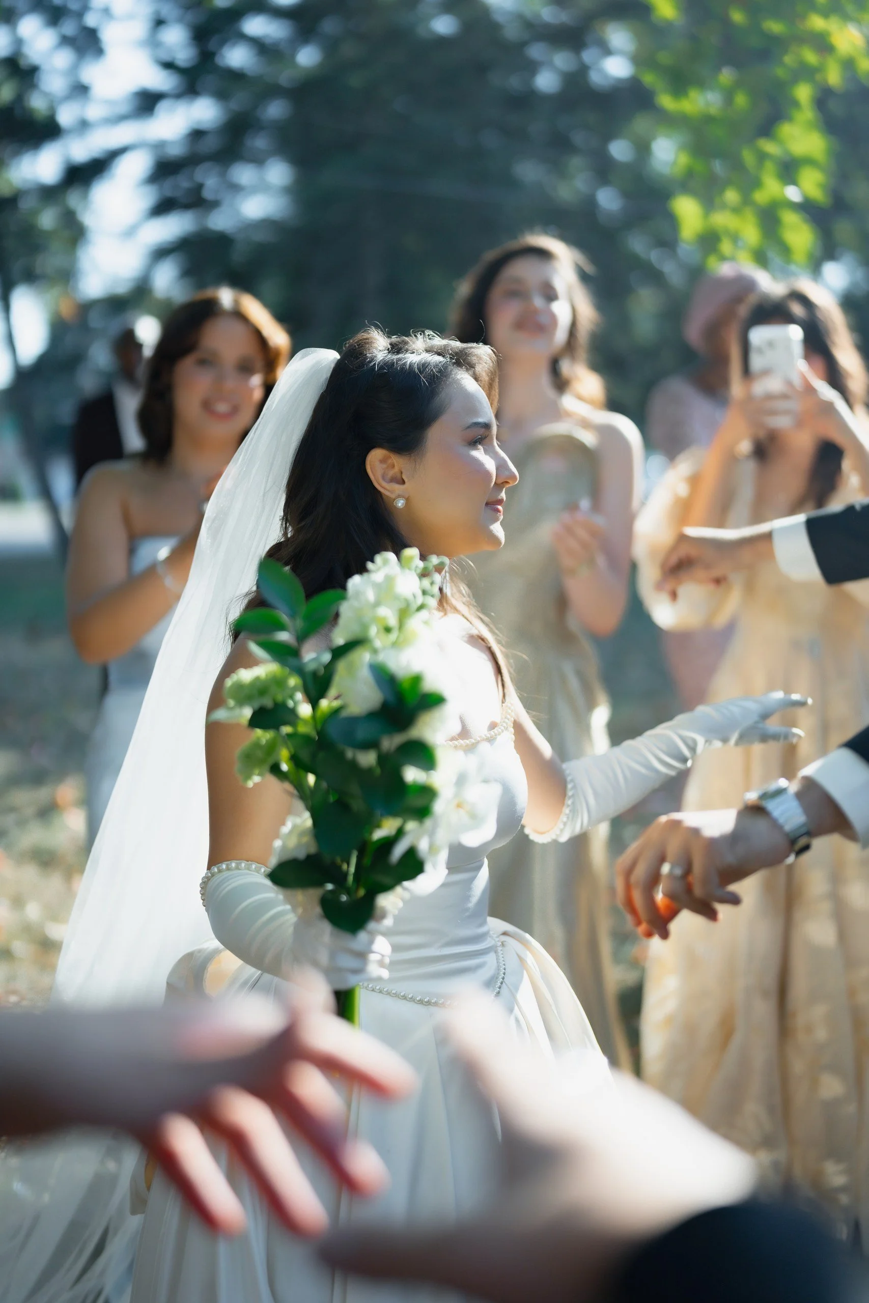 A bride holding a bouquet of white flowers during a wedding ceremony outdoors, surrounded by women in elegant dresses.