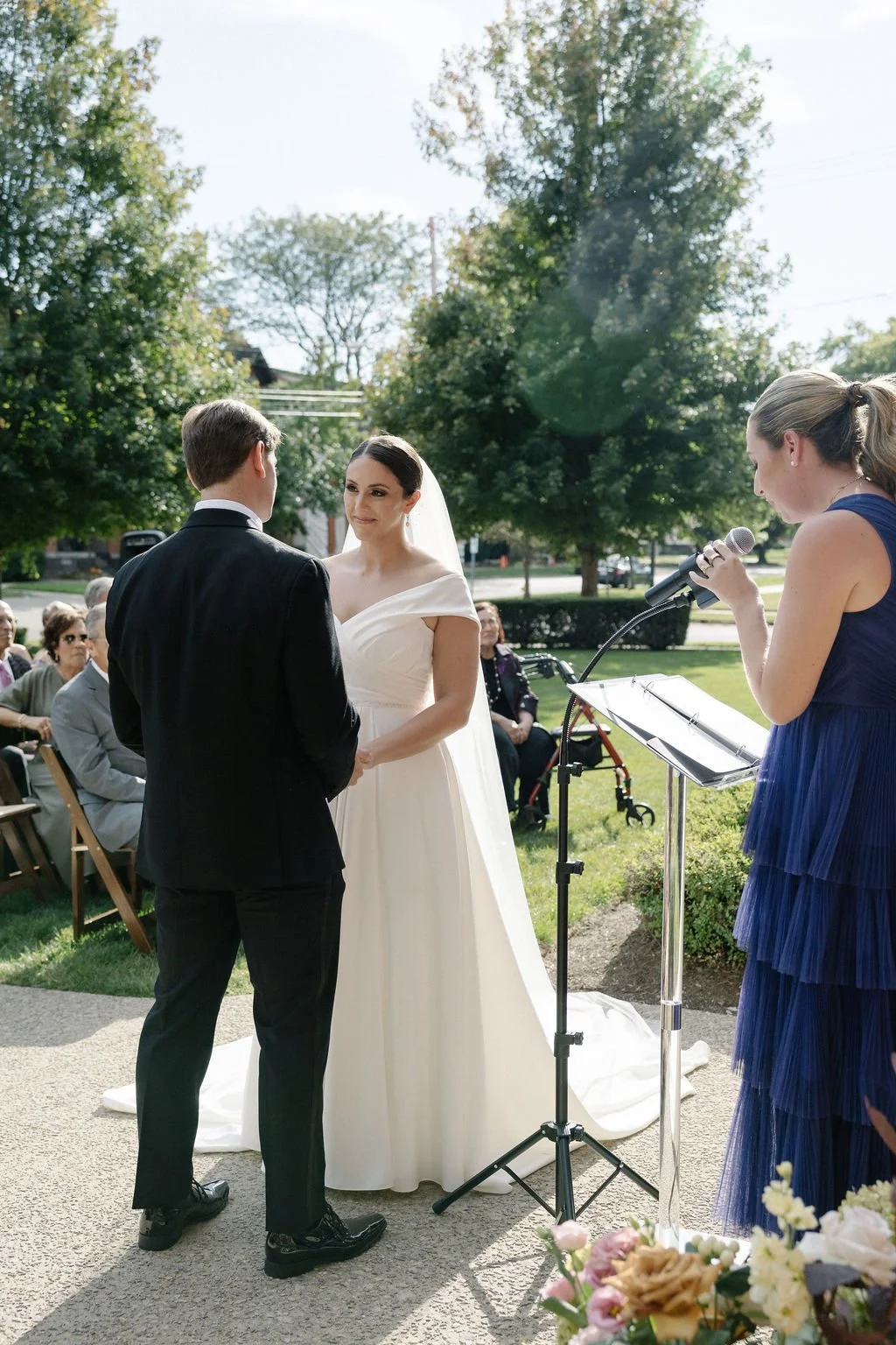 A couple gets married outdoors, with a woman reading vows into a microphone as friends and family look on.