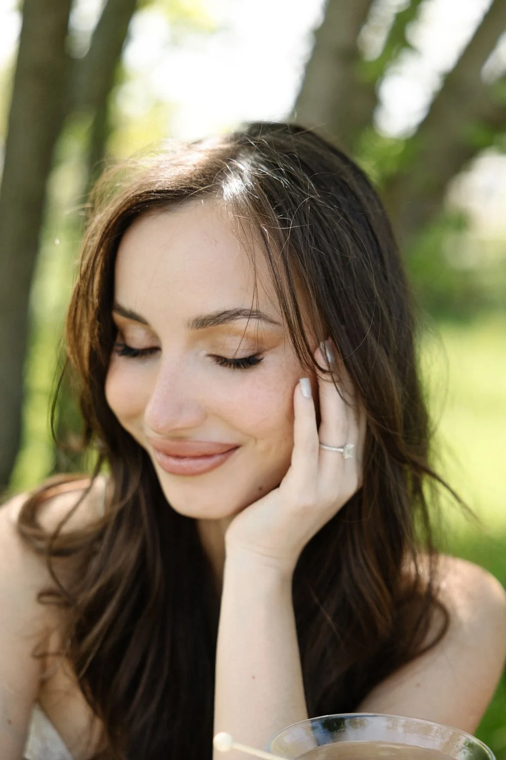 Close-up of a young woman with long brown hair, smiling with her eyes closed, outdoors in natural sunlight.