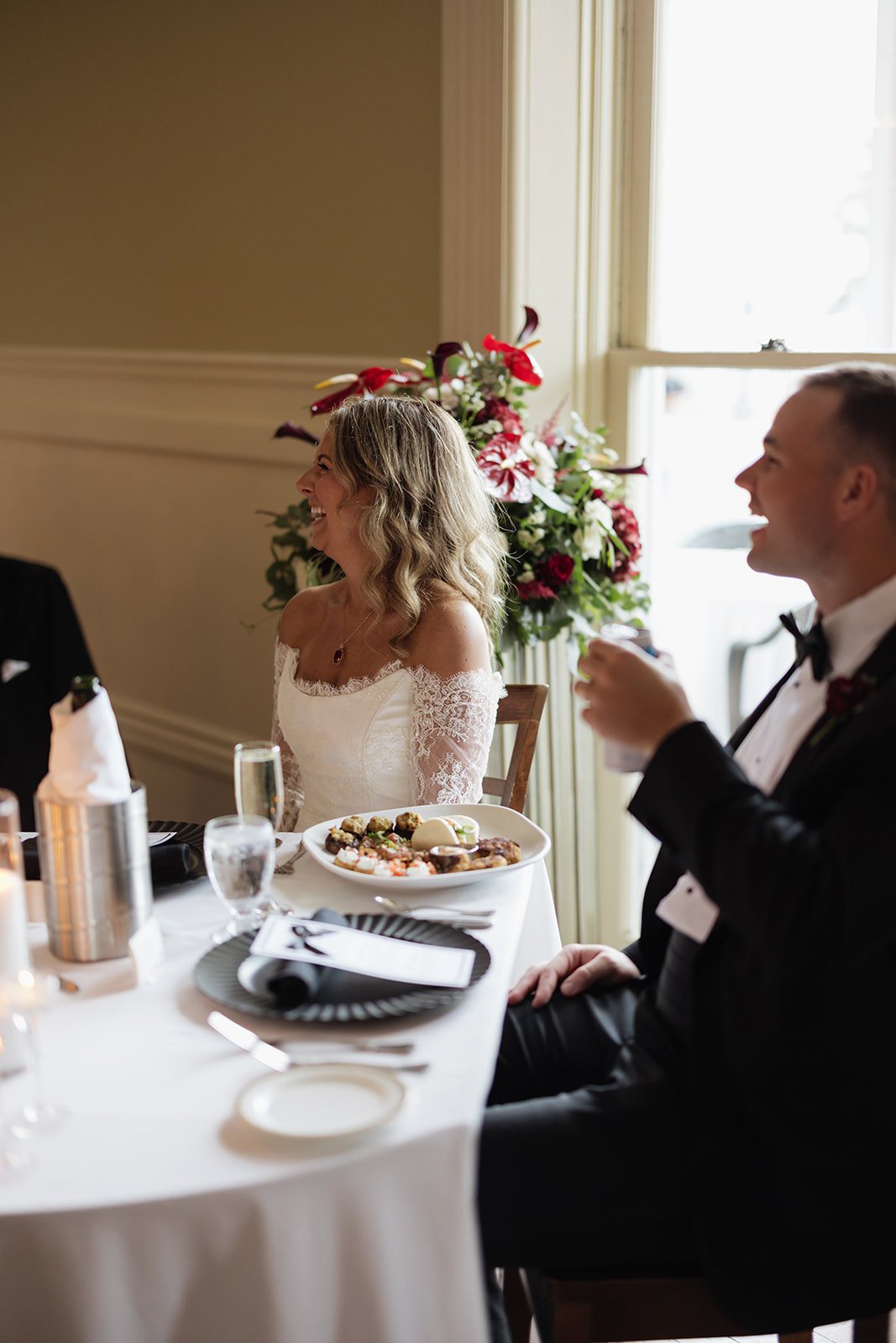 A bride and groom sitting at a decorated table during a wedding reception, smiling and laughing indoors with a large floral arrangement in the background.