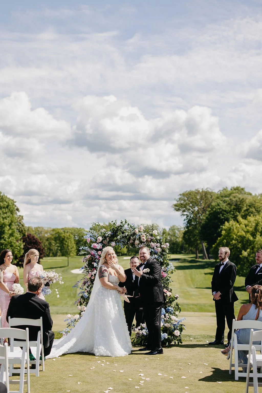 Wedding ceremony outdoors on a grassy field with a floral arch, a bride and groom exchanging vows, surrounded by bridesmaids and groomsmen, with trees and a cloudy sky in the background.