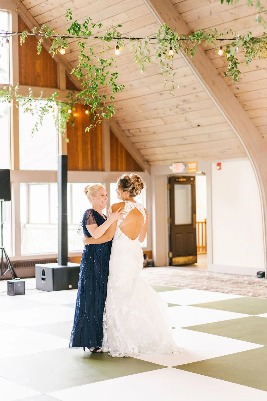 A bride in a white lace wedding dress dancing with a woman in a dark blue dress inside a rustic wood-paneled venue decorated with hanging greenery and string lights.