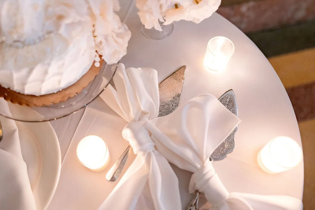 Elegant table setting with a cake on a cake stand, white napkins tied with ribbons, and small lit candles on a white tablecloth