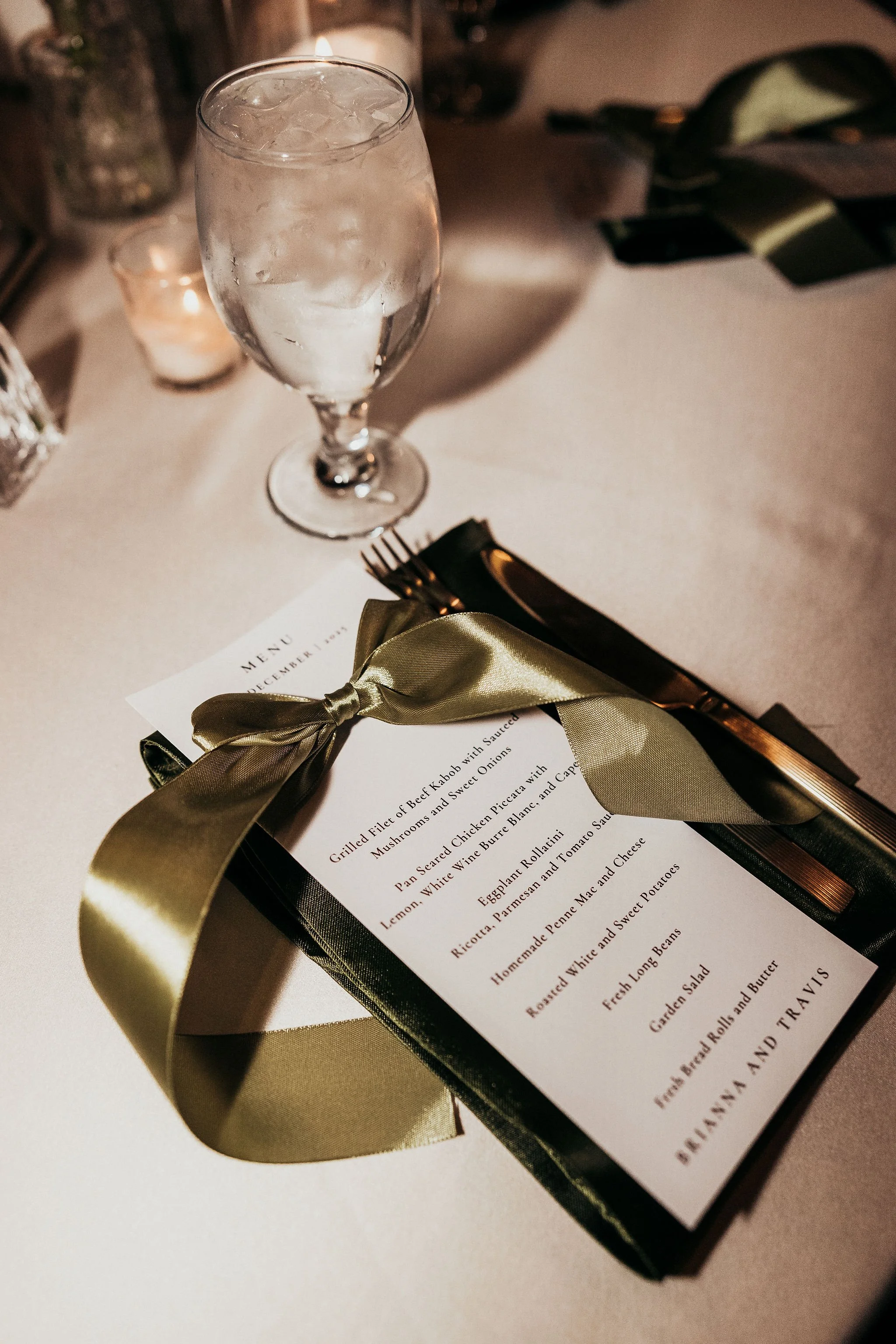 Elegant table setting with a glass of sparkling water, a gift-wrapped item with a gold ribbon, and a candle in a glass holder, alongside a menu card.