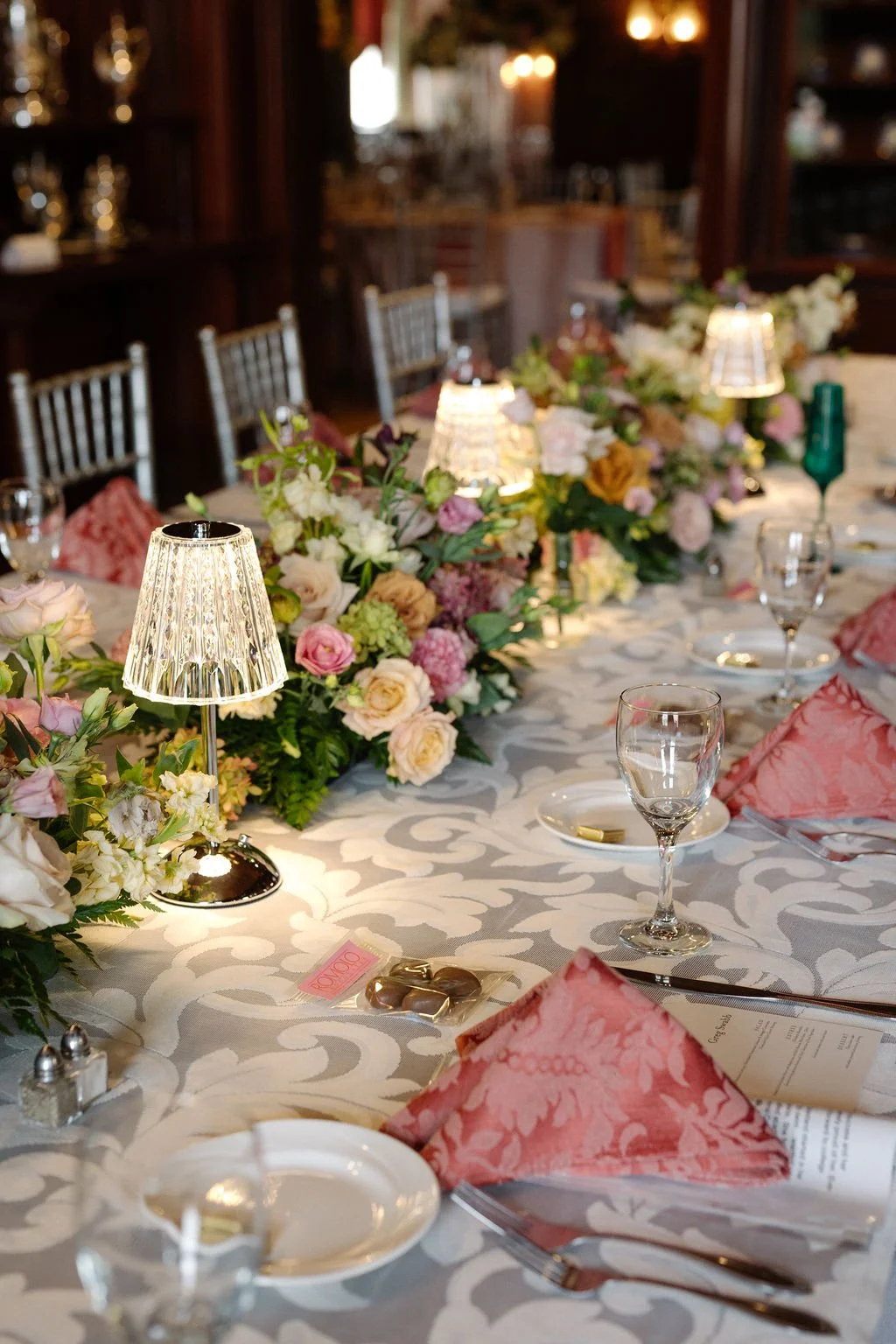 A decorated banquet table with floral arrangements, small lamps, pink napkins, and glassware in a dimly lit elegant setting.