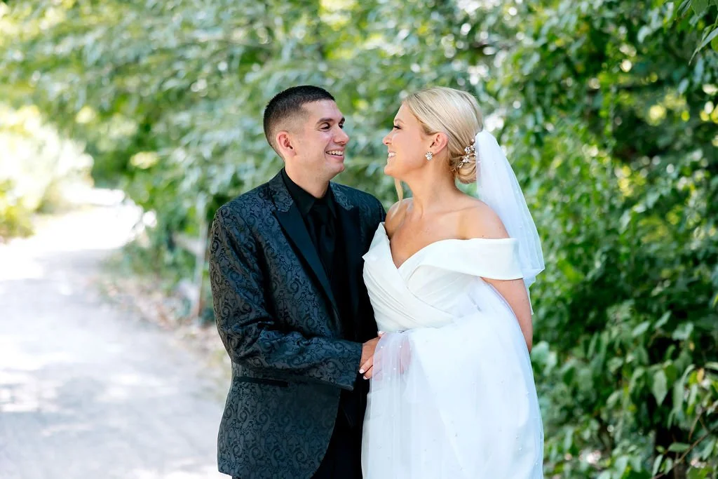 A bride and groom stand together outdoors surrounded by green foliage, smiling at each other.