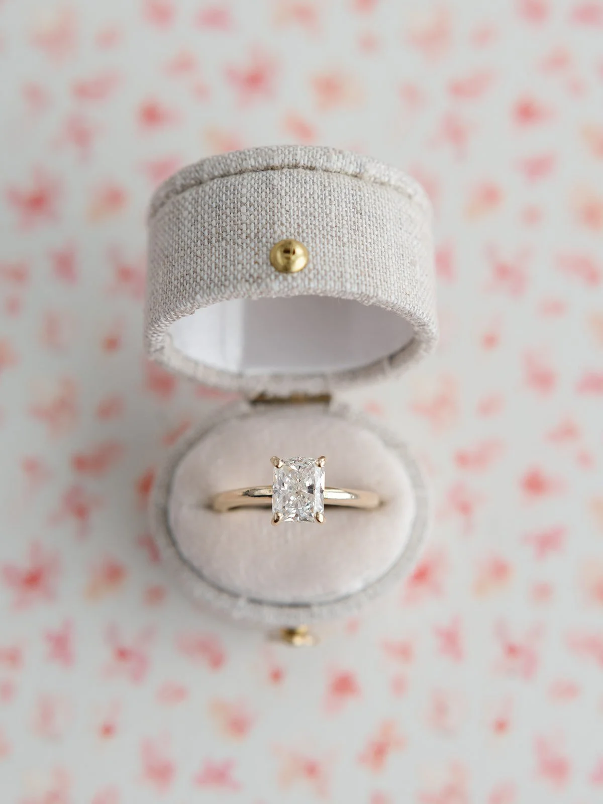A diamond engagement ring in a white ring box with a rectangular cut stone, open on a pink and red patterned background.