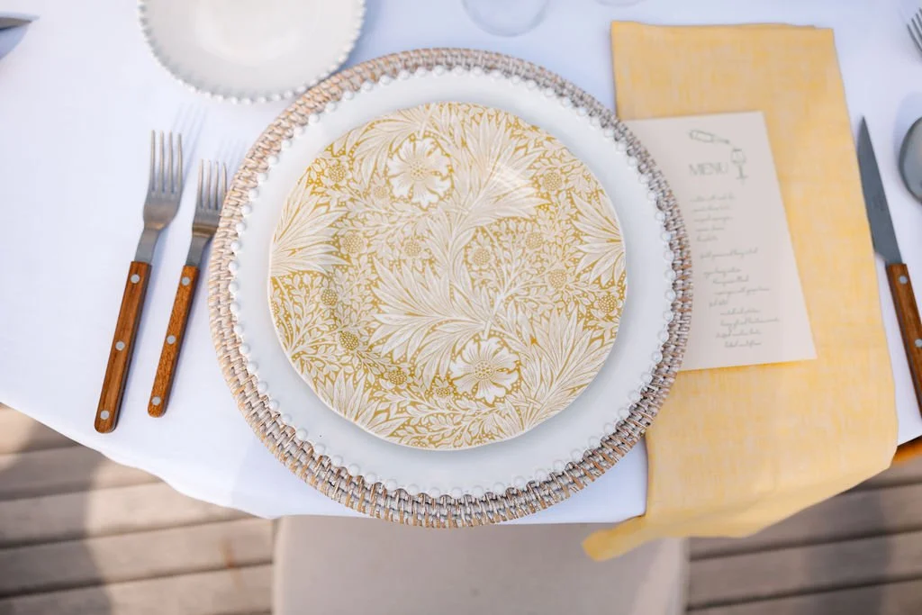 A table setting with a patterned white and gold floral plate, a layered charger plate, two forks with wooden handles, a yellow napkin, silverware, a glass, and a menu on a white tablecloth.