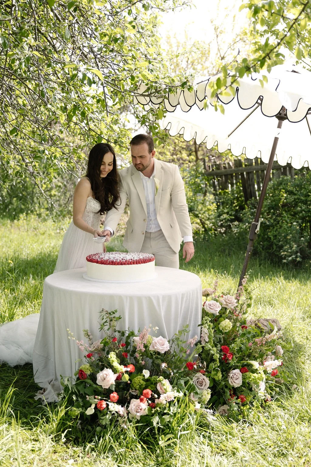 A bride and groom cutting a wedding cake outdoors under a white parasol. They are standing at a table with a white tablecloth, surrounded by floral arrangements, in a green garden with trees and a wooden fence.