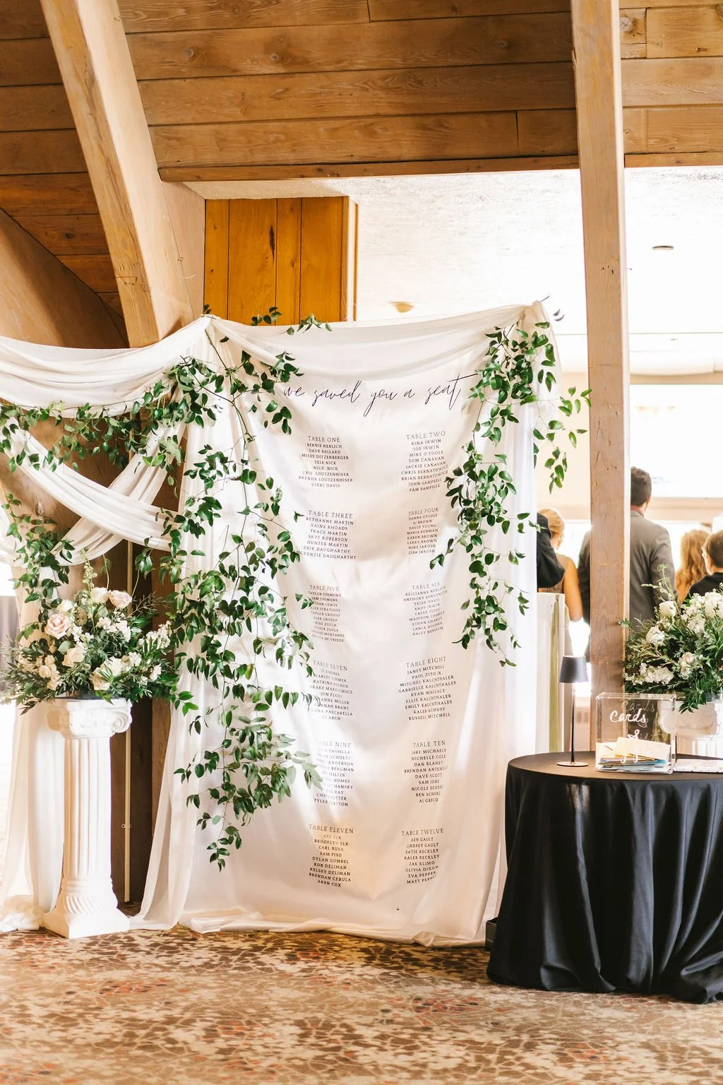 Wedding seating chart display on white fabric with green foliage, floral arrangements, and table number listings, set against a wooden wall at the reception venue.