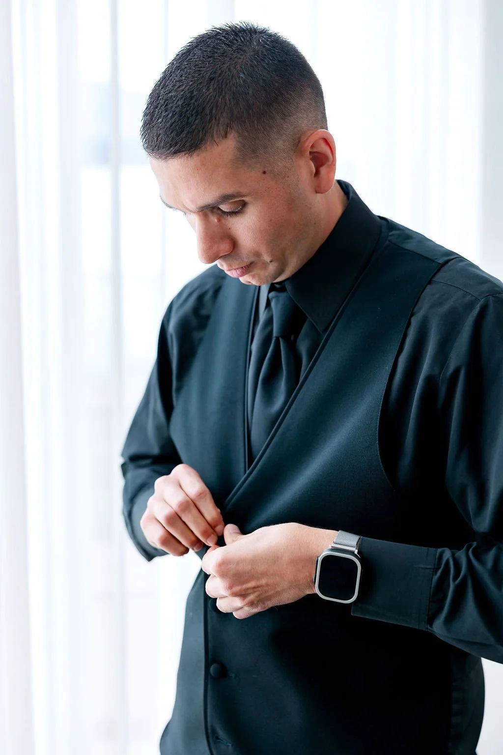 Man in black suit adjusting his jacket in front of a white curtain background, wearing a smartwatch on his left wrist.