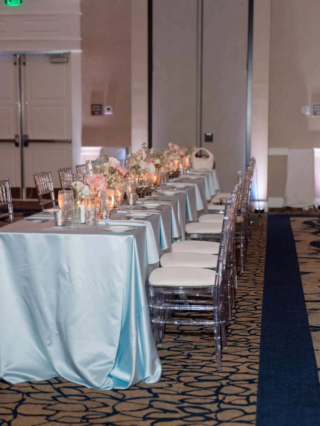 A long banquet table decorated with pink and white flowers, candles, and tableware in a formal event room with patterned carpet and neutral walls.